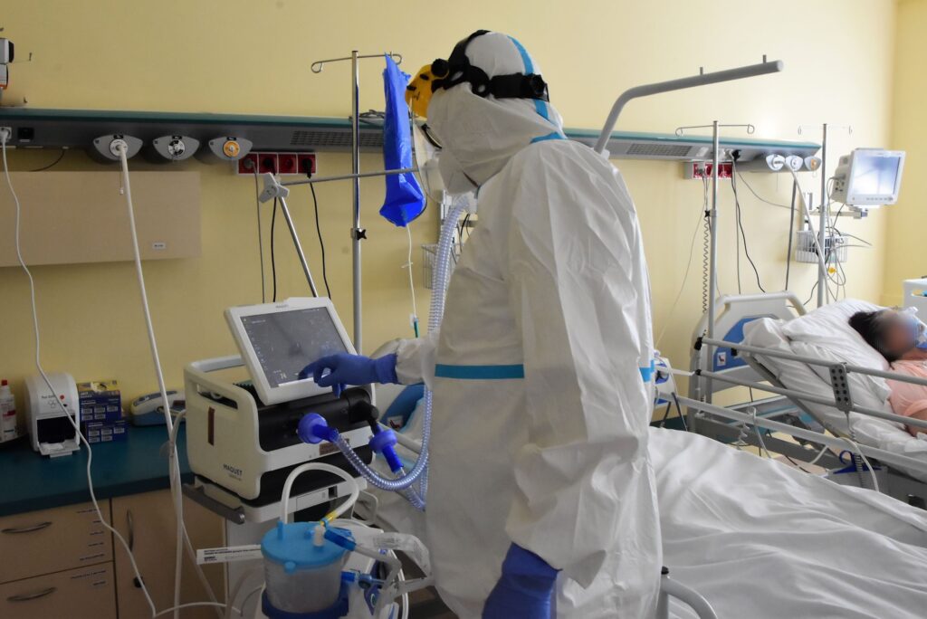lekarz w kombinezonie ochronnym sprawdza ustawienia na monitorze respiratora, obok leży zaintubowany pacjent